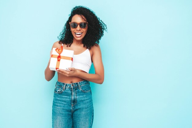 Schöne schwarze Frau mit Afro-Locken-Frisur Lächelndes Modell in weißer Sommerkleidung gekleidet Sexy sorglose Frau posiert in der Nähe der blauen Wand im Studio Gebräunt und fröhlich Holding-Geschenkbox Isoliert