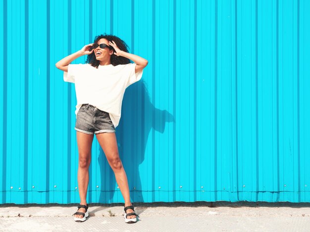 Schöne schwarze Frau mit Afro-Locken-FrisurLächelndes Hipster-Modell in weißem T-Shirt Sexy sorglose Frau posiert auf der Straße in der Nähe der blauen Wand mit Sonnenbrille Fröhlich und glücklich