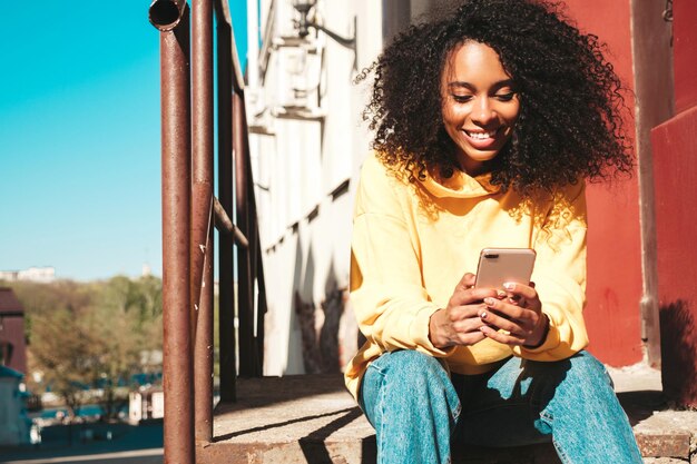 Schöne schwarze Frau mit Afro-Locken-FrisurLächelndes Modell in gelbem Hoodie Sexy sorglose Frau, die mit Sonnenbrille auf dem Straßenhintergrund posiert Blick auf den Smartphone-Bildschirm mit Apps