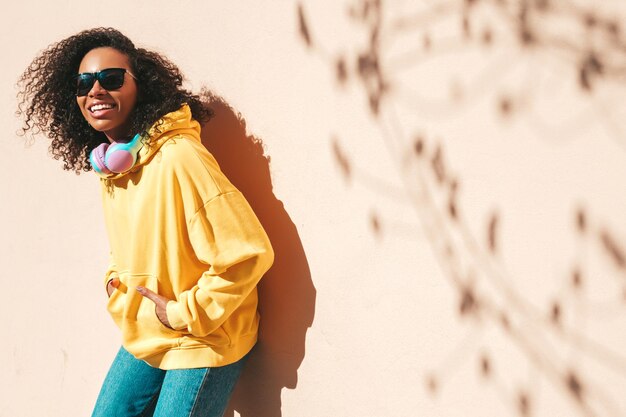 Schöne schwarze Frau mit Afro-Locken-FrisurLächelndes Modell in gelbem HoodieSexy sorglose Frau, die Musik in drahtlosen Kopfhörern hörtPosing auf Straßenhintergrund in der Nähe einer Wand mit Sonnenbrille