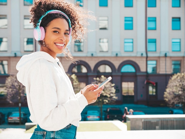 Schöne schwarze Frau mit Afro-Locken-FrisurLächelndes Modell in weißem HoodieSexy sorglose Frau, die das Hören von Musik in drahtlosen Kopfhörern genießtPosing on street background at sunsetHolds phone