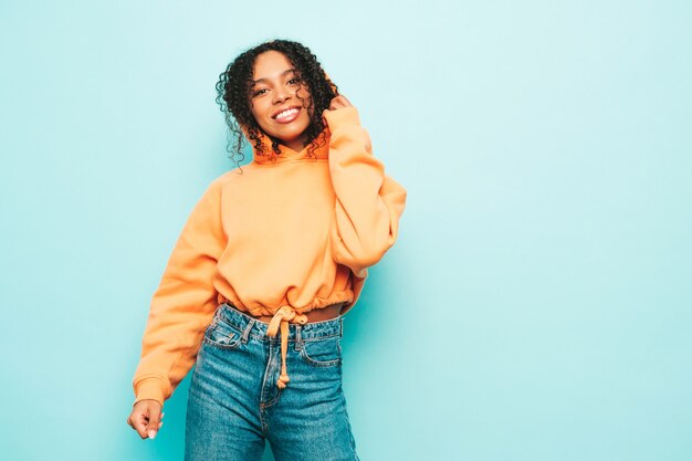 Schöne schwarze Frau mit Afrolockenfrisur. Lächelndes Model in orangefarbenem Hoodie und trendiger Jeanskleidung