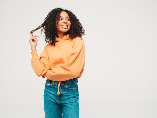 Schöne schwarze Frau mit Afrolockenfrisur. Lächelndes Model in orangefarbenem Hoodie und trendiger Jeanskleidung