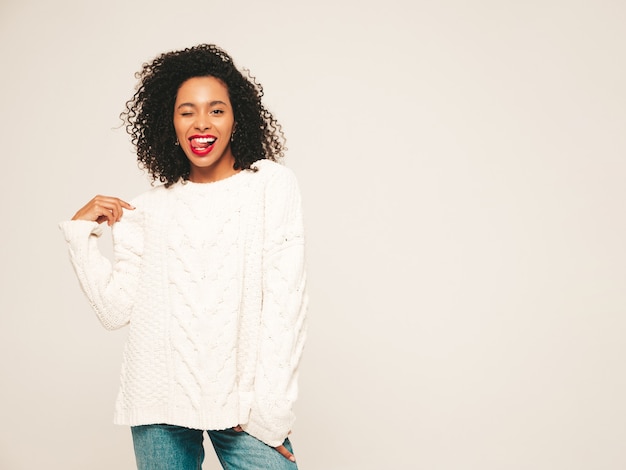 Schöne schwarze Frau mit Afrolockenfrisur. Lächelndes Modell in der weißen Winterstrickjacke und in der Jeanskleidung.