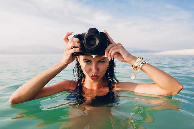 Schöne sexy Frau, gebräunte Haut, schwarzer Bikini-Badeanzug, im blauen Wasser stehend, digitale Fotokamera haltend, heißer, tropischer Sommerurlaub, Modetrend, flirtend, nass