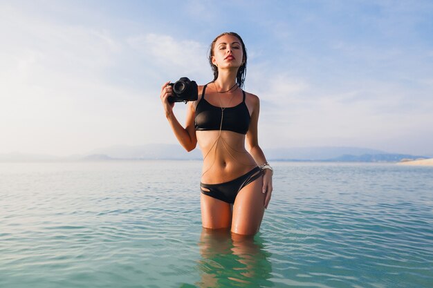 Schöne sexy Frau, perfekter schlanker Körper, gebräunte Haut, schwarzer Bikini-Badeanzug, im blauen Wasser stehend, digitale Fotokamera haltend, heißer, tropischer Sommerurlaub, Modetrend, Taille, Bauch, Hüften