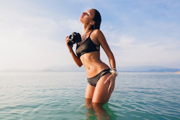 Schöne sexy Frau, perfekter schlanker Körper, gebräunte Haut, schwarzer Bikini-Badeanzug, im blauen Wasser stehend, digitale Fotokamera haltend, heißer, tropischer Sommerurlaub, Modetrend, Taille, Bauch, Hüften