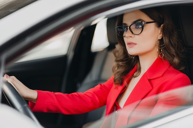 Schöne sexy reiche Geschäftsfrau im roten Anzug, der im weißen Auto fährt, Brille tragend, Geschäftsdamenstil