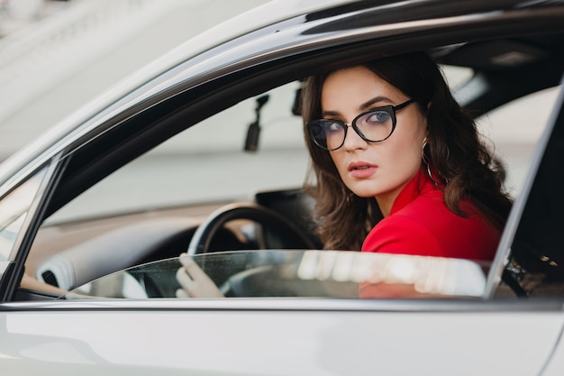 Schöne sexy reiche Geschäftsfrau im roten Anzug, der im weißen Auto sitzt und Brille trägt