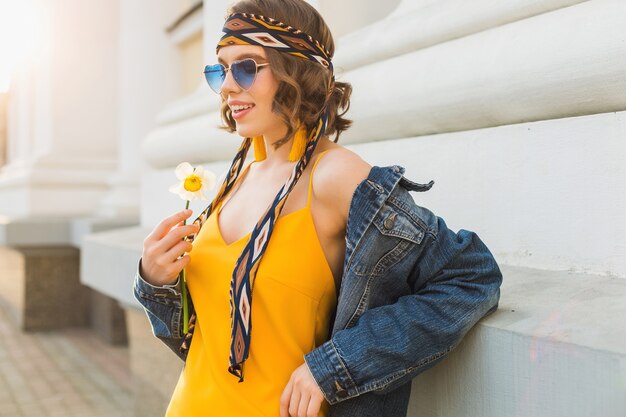 Schöne sexy stilvolle Frau im gelben stilvollen Kleid, das Jeansjacke, trendiges Outfit, Frühlingssommer-Modetrend, sonnige, blaue Sonnenbrille, Straßenmode, Hipster-Stil, modische Accessoires trägt