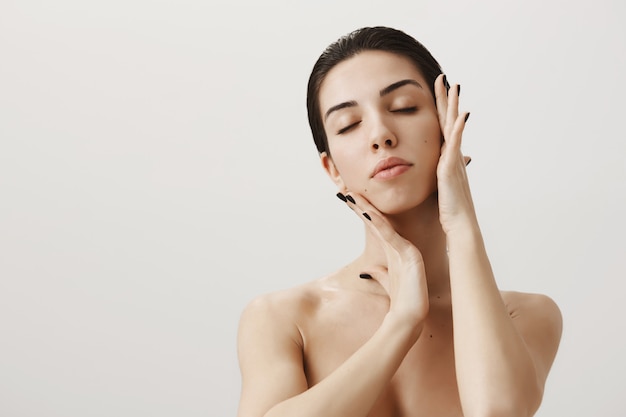 Schöne sinnliche Frau, die nackt mit geschlossenen Augen steht und Gesicht sanft berührt, um Hautpflegeprodukt aufzutragen