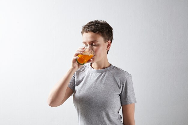 Schöne sportliche Frau mit gesund perfekter Haut im einfachen grauen T-Shirt, das träumerisch ihren frischen organischen Karottenorangensaft trinkt