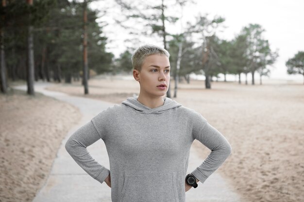 Schöne sportliche junge kurzhaarige blonde Frau, die müden Blick hat, Atem nach Morgenlauf holt, Hände auf ihrer Taille hält, auf gepflastertem Weg gegen Sandstrand und Kiefernhintergrund aufwirft