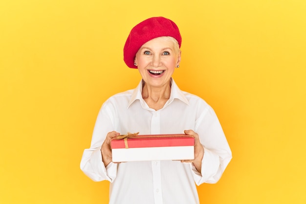 Schöne stilvolle Dame im Ruhestand in weißem Hemd und roter Baskenmütze, aufgeregt mit angenehmer Überraschung, die Schachtel der Süßigkeiten am internationalen Frauentag empfangend, den Mund mit Aufregung öffnend