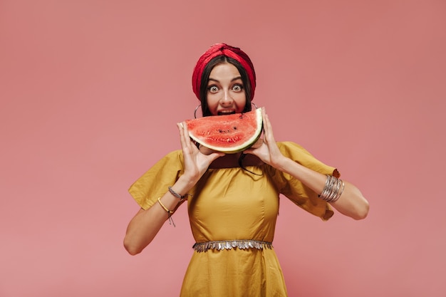 Schöne stilvolle Dame in rotem Bandana, modernen Armbändern und gelbem hellem Kleid, das in die Kamera schaut und Wassermelone isst