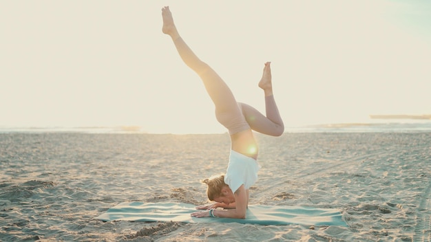 Schöne stilvolle Frau, die Yoga-Pose-Handstand mit Spagat am leeren Strand macht Junge, atemberaubende Yogalehrerin, die Yoga auf der Matte praktiziert