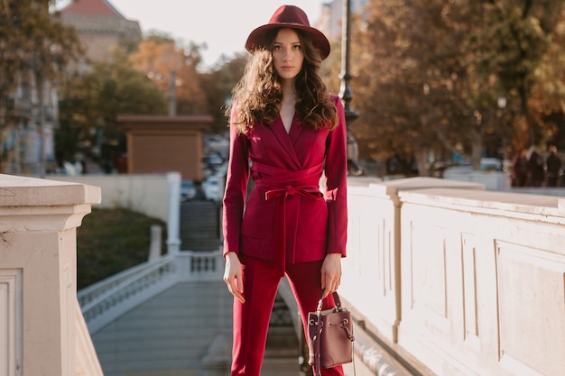 Schöne stilvolle Frau im lila Anzug, der in der Stadtstraße, Frühlingssommer-Herbstsaison-Modetrend trägt Hut trägt und Geldbörse hält