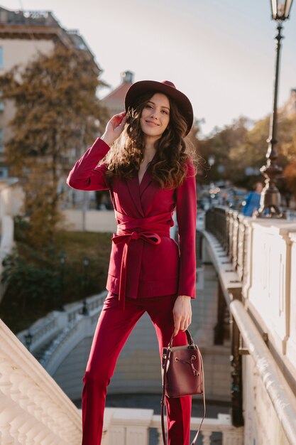 Schöne stilvolle Frau im lila Anzug, der in der Stadtstraße, Frühlingssommer-Herbstsaison-Modetrend trägt Hut trägt und Geldbörse hält