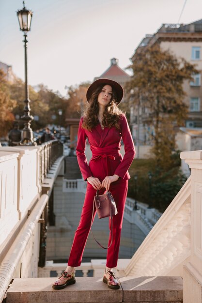 Schöne stilvolle Frau im lila Anzug, der in der Stadtstraße, Frühlingssommer-Herbstsaison-Modetrend trägt Hut trägt und Geldbörse hält