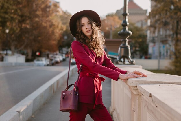 Schöne stilvolle Frau im lila Anzug, der in der Stadtstraße, Frühlingssommer-Herbstsaison-Modetrend trägt Hut trägt und Geldbörse hält