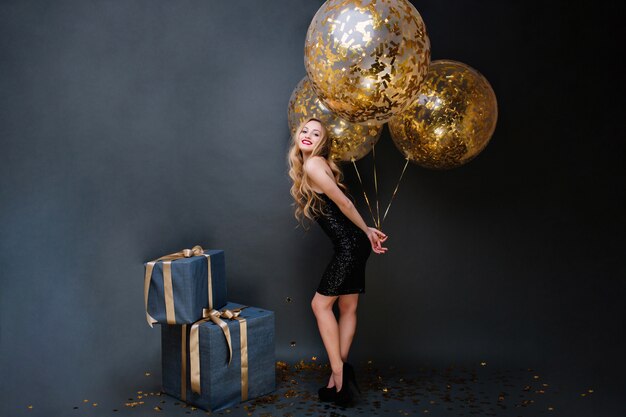 Schöne stilvolle junge Frau auf Absätzen, langes lockiges blondes, schwarzes Luxuskleid mit großen Luftballons voll mit goldenen Lametta. Geschenke, Geburtstagsfeier, Feiern, Lächeln.