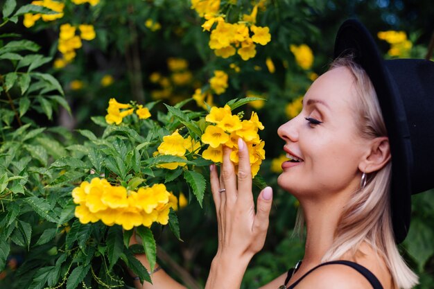 Schöne stilvolle kaukasische glückliche Frau im schwarzen Kleid und im klassischen Hut im Park, umgeben von gelben thailändischen Blumen