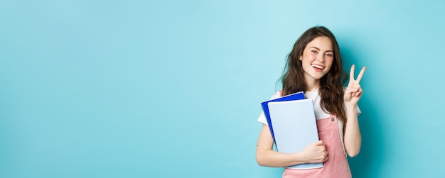 Schöne Studentin, die Vsign zeigt und glücklich lächelt, wenn sie Notizbücher mit Lernmaterial hält