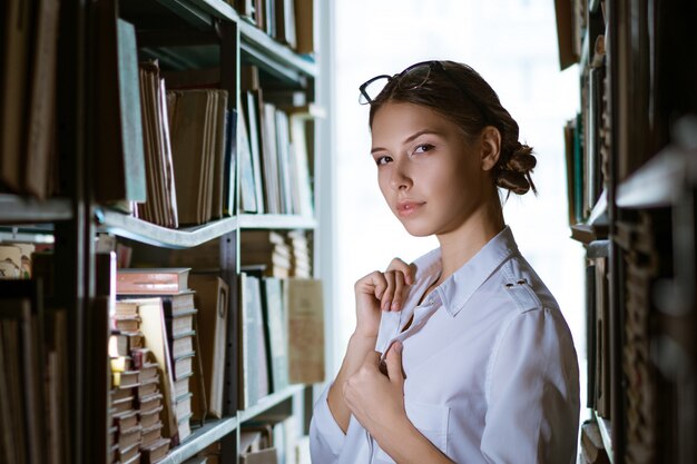 Schöne Studentin in einem weißen Hemd steht zwischen den Reihen in der Bibliothek