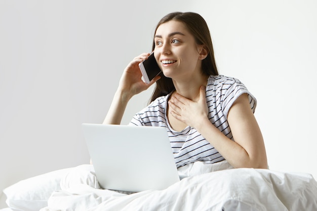 Schöne Studentin in gestreiften Pyjamas, die Morgen im Bett verbringen, Internet mit Laptop surfen und auf Handy sprechen