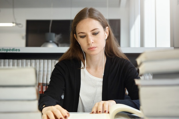 Kostenloses Foto schöne teenagerfrau mit langen brünetten haaren, die uniform tragen, die lehrbuch oder handbuch studiert und ihre lieblingsmusik mit kopfhörern beim sitzen in der schulbibliothek hört