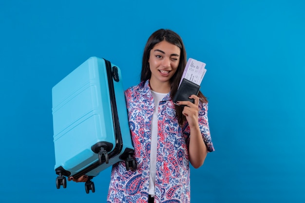 Schöne Touristenfrau, die Reisekoffer und Reisepass mit Eintrittskarten mit Lächeln auf Gesicht hält, das glückliches und positives Reisekonzept über blauem Raum zwinkert