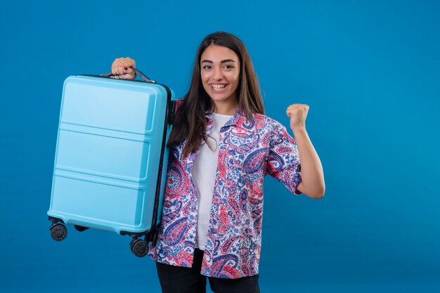 Schöne touristische Frau mit dem Reisekoffer, der aufgeregt schaut und sich über ihren Erfolg und Sieg freut, die ihre Fäuste mit der Freude ballen, glücklich, ihr Ziel und ihre Ziele zu erreichen, die über isoliertem blauem Hintergrund stehen