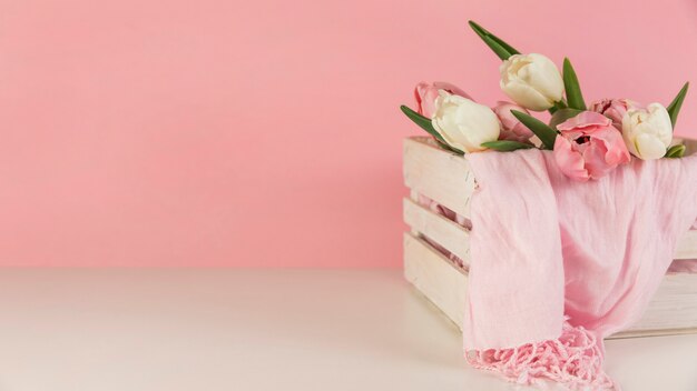 Schöne Tulpen und Schal in der hölzernen Kiste auf weißem Schreibtisch gegen rosa Hintergrund