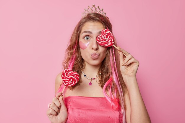 Schöne überraschte Frau bedeckt das Auge mit süßen leckeren Süßigkeiten hält die Lippen gerundet trägt Krone und Kleid trägt Schönheitsflecken unter den Augen für die Hautpflege auf, die über rosa Hintergrund isoliert sind Süßwaren