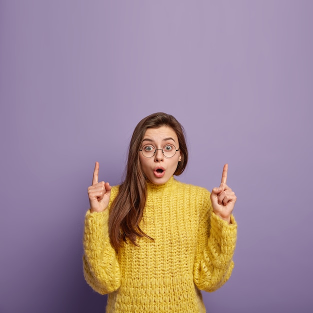 Schöne überraschte Frau hat erschrockenen Blick, zeigt auf etwas Unglaubliches, trägt runde Brille und gelben Strickpullover, zeigt einige Artikel im Laden, isoliert über lila Wand