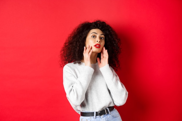 Schöne und stilvolle lockige Frau mit roten Lippen, die das perfekte junge Gesicht berührt und sinnlich in die Kamera schaut, vor rotem Hintergrund stehend