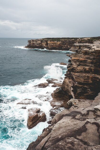 Schöne vertikale Aufnahme einer großen Klippe neben blauem Wasser an einem trüben Tag