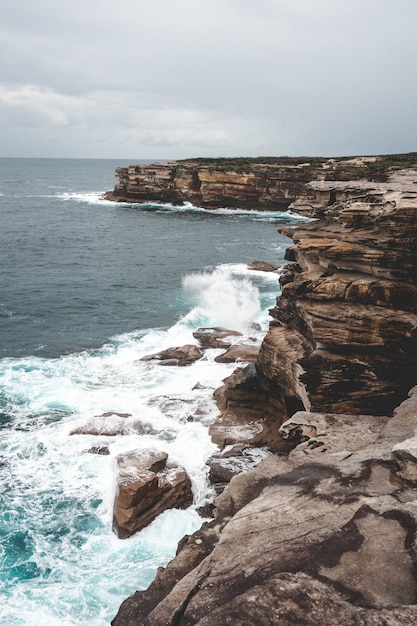 Schöne vertikale Aufnahme einer großen Klippe neben blauem Wasser an einem trüben Tag