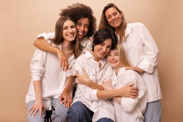Schöne, vielfältige junge Damen in Jeans und weißen Hemden schauen auf beigem Hintergrund in die Kamera Frauentagskonzept