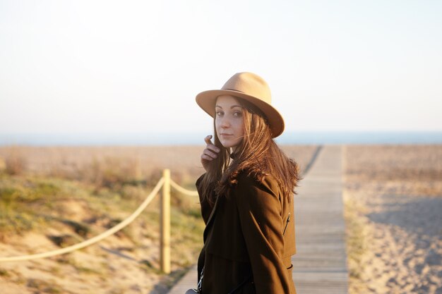 Schöne zarte junge europäische Frau, die stilvollen Hut und Mantel trägt, die entlang Promenade gehen
