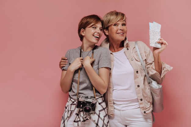Schöne zwei Frauen mit stilvoller kurzer Frisur in der modernen Kleidung, die zwei Karten und Kamera auf rosa Hintergrund umarmt, lacht und hält.