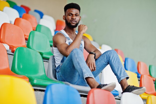 Schöner afrikanisch-amerikanischer Mann in Jeans-Overalls posierte auf farbigen Stühlen im Stadion Modisches Porträt eines schwarzen Mannes