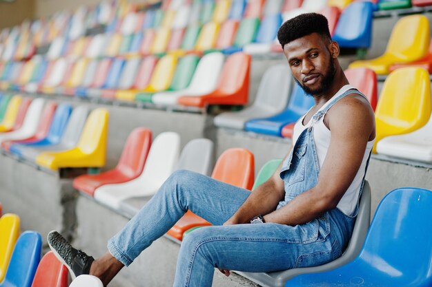 Schöner afrikanisch-amerikanischer Mann in Jeans-Overalls posierte auf farbigen Stühlen im Stadion Modisches Porträt eines schwarzen Mannes