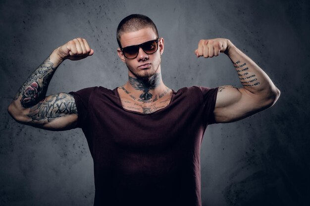 Schöner, athletischer Mann mit Sonnenbrille mit Tätowierungen an Armen und Hals, der in einem Studio vor grauem künstlerischem Hintergrund posiert.