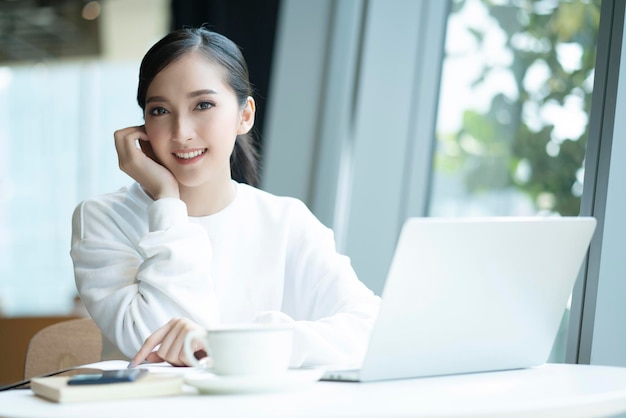 Schöner attraktiver asiatischer weiblicher Frischelächeln-fröhlicher Morgen, der mit Laptopkaffeearbeit von zu Hause aus mit Glück arbeitet, genießt Blick auf den Garten asiatische junge Frau mit Fensterlicht-Gartenhintergrund