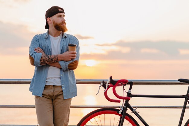 Schöner bärtiger Mann des ernsthaften Hipster-Stils, der Jeanshemd und Mütze mit Fahrrad im Morgensonnenaufgang durch das Meer trägt, das Kaffee trinkt, gesunder Reisender des aktiven Lebensstils