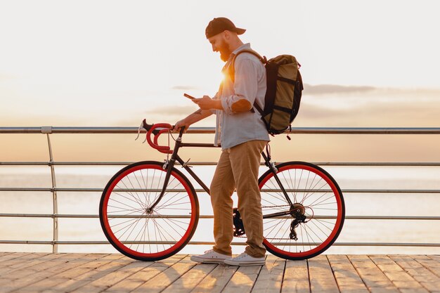 Schöner bärtiger Mann des Hipster-Stils, der mit Rucksack auf Fahrrad unter Verwendung des Telefons im Morgensonnenaufgang durch das Meer reist, gesunder aktiver Lebensstil