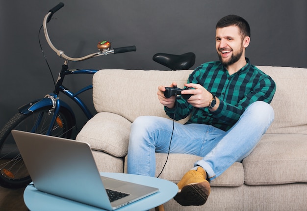 Schöner bärtiger Mann des jungen Hipsters, der zu Hause auf der Couch sitzt und Videospiel auf Notizbuch spielt