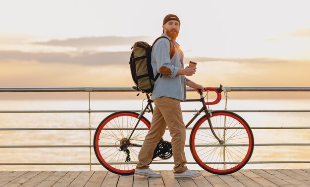 Schöner bärtiger Mann im Hipster-Stil mit Rucksack, der Jeanshemd und Mütze mit Fahrrad im Morgensonnenaufgang durch das Meer trägt, das Kaffee trinkt, gesunder aktiver Lebensstil reisender Rucksacktourist