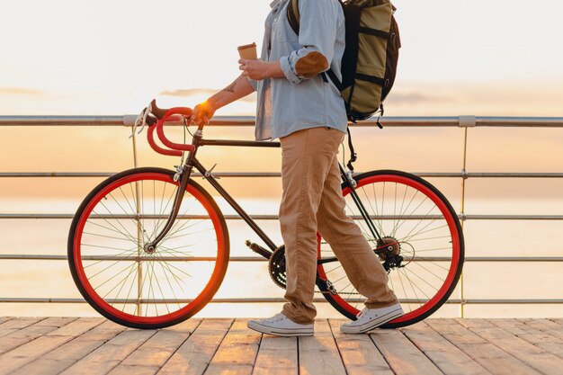 Schöner bärtiger Mann im Hipster-Stil mit Rucksack, der Jeanshemd und Mütze mit Fahrrad im Morgensonnenaufgang durch das Meer trägt, das Kaffee trinkt, gesunder aktiver Lebensstil reisender Rucksacktourist